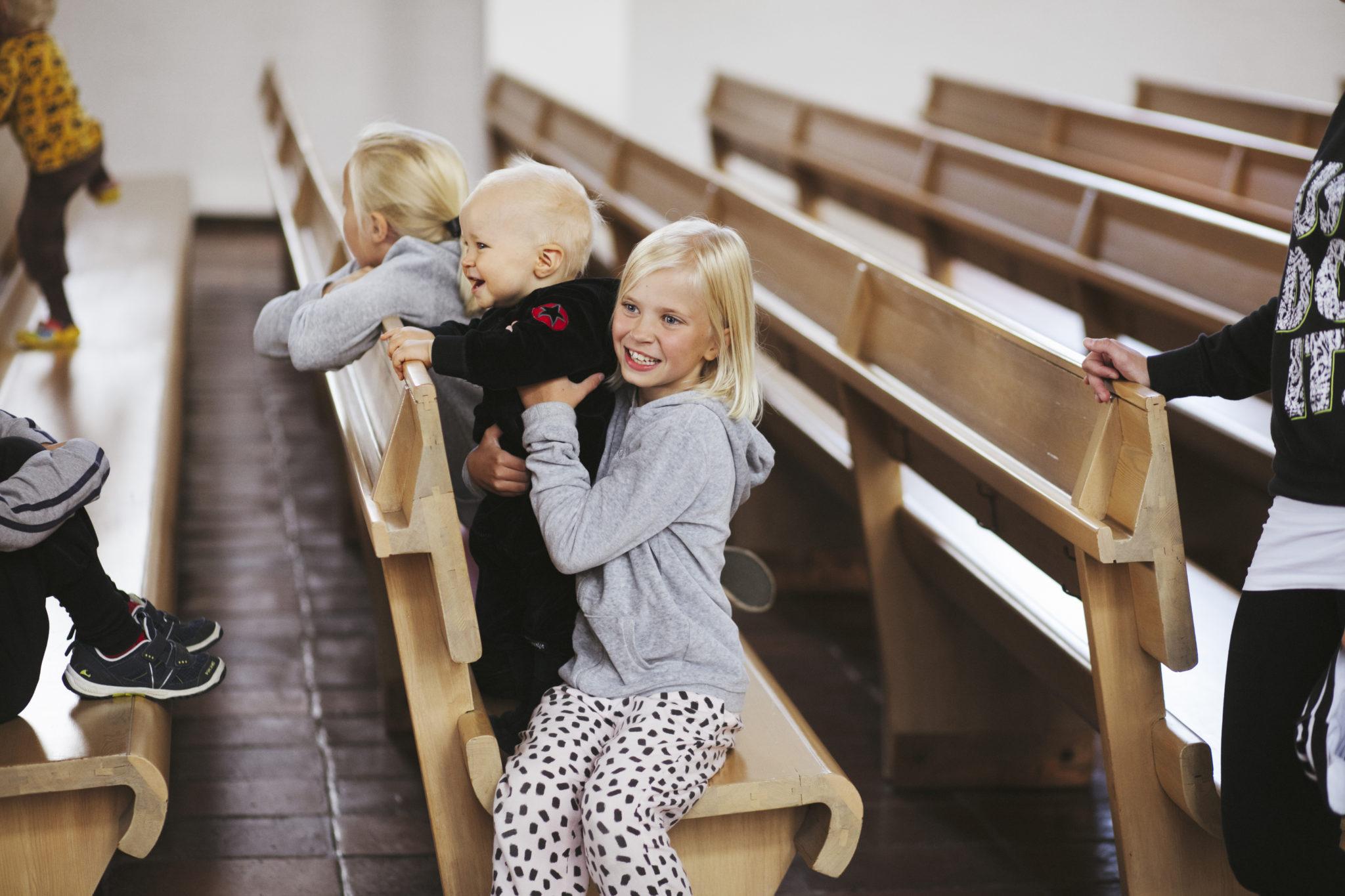Lapset istuvat Lakeuden Ristin kirkon penkillä. Sisko pitää veljeä sylissä nauraen.