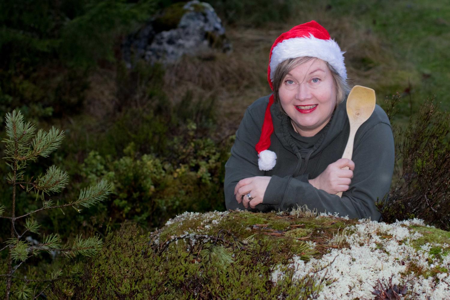 Kokki Nanna Rintala nojaa tonttulakki päässään ja puinen kauha kädessään sammaleiselle kivelle metsässä.