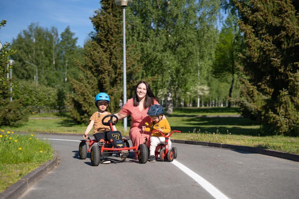 Lapset ja äiti poseeraavat Liikenne ja fiilispuistossa. Lapset istuvat polkuautojen kyydissä.