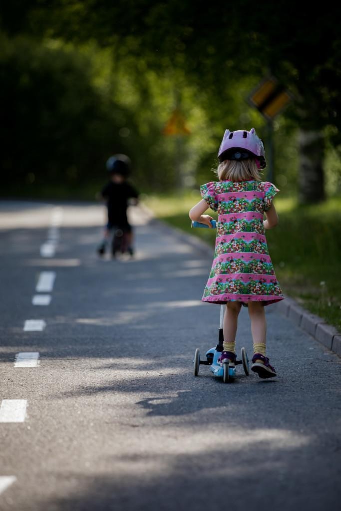 Lapsista toinen ajaa pienellä pyörällä, jossa apurenkaat ja toinen potkulaudalla liikennepuistossa.