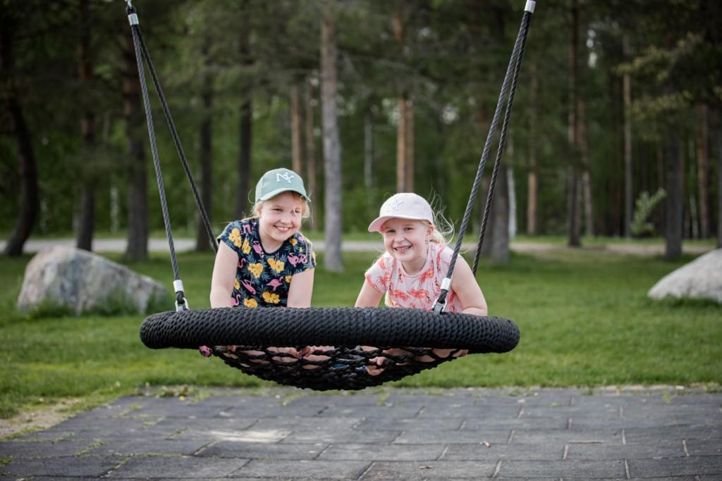Lapset keinumassa korikeinussa hymyillen Kyrkösjärven maisemissa.