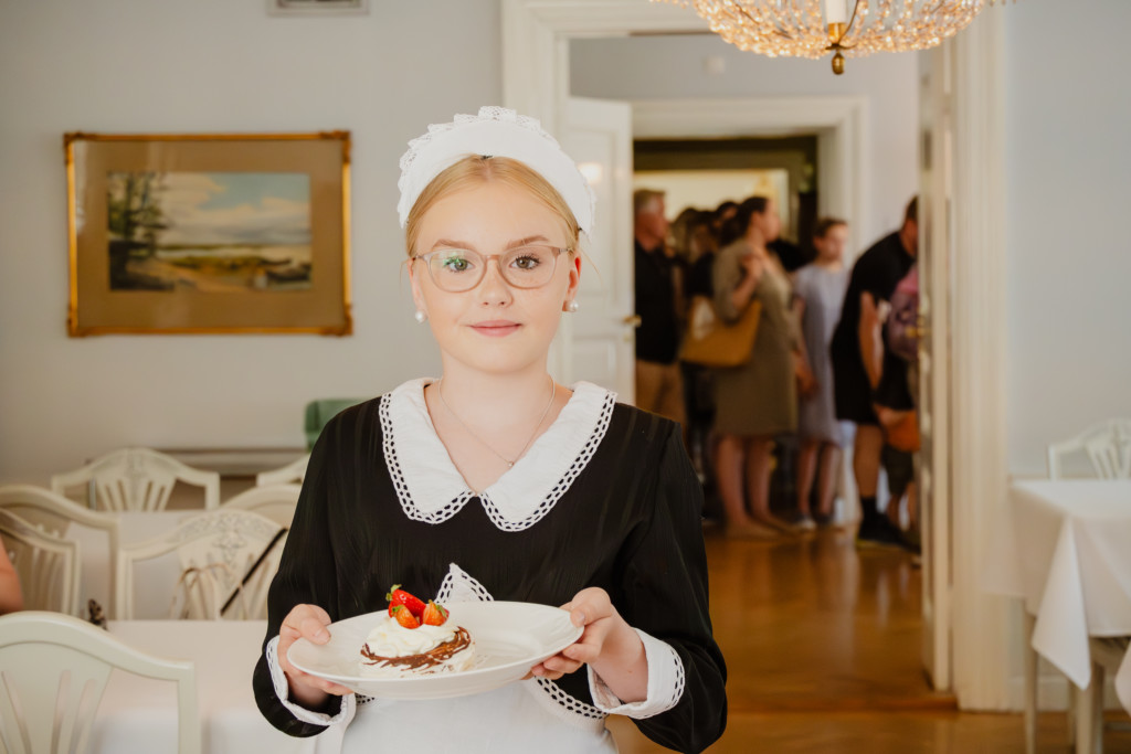 Nuori nainen esittelee lautasella olevaa kakkupalaa, hän on pukeutunut historialliseen mustavalkoiseen tarjoilijan asuun