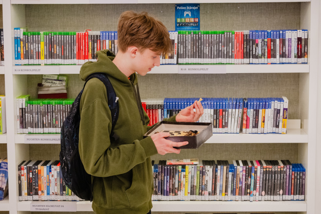 Poika katselee pelejä kirjaston hyllyllä pitäen shakkilautaa käsissään